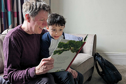 James on a sofa in a comfortable room, reading a book with a young child. Both James and the child look really excited at what they're reading.