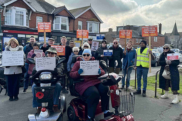James MacCleary in a crowd of local residents holding placards protesting about uncontrolled parking in Polegate. At the front are two people in mobility scooters with handwritten signs saying "Please don't park across dropped kerbs". Several others are holding printed signs saying "Uncontrolled parking is destroying our town".